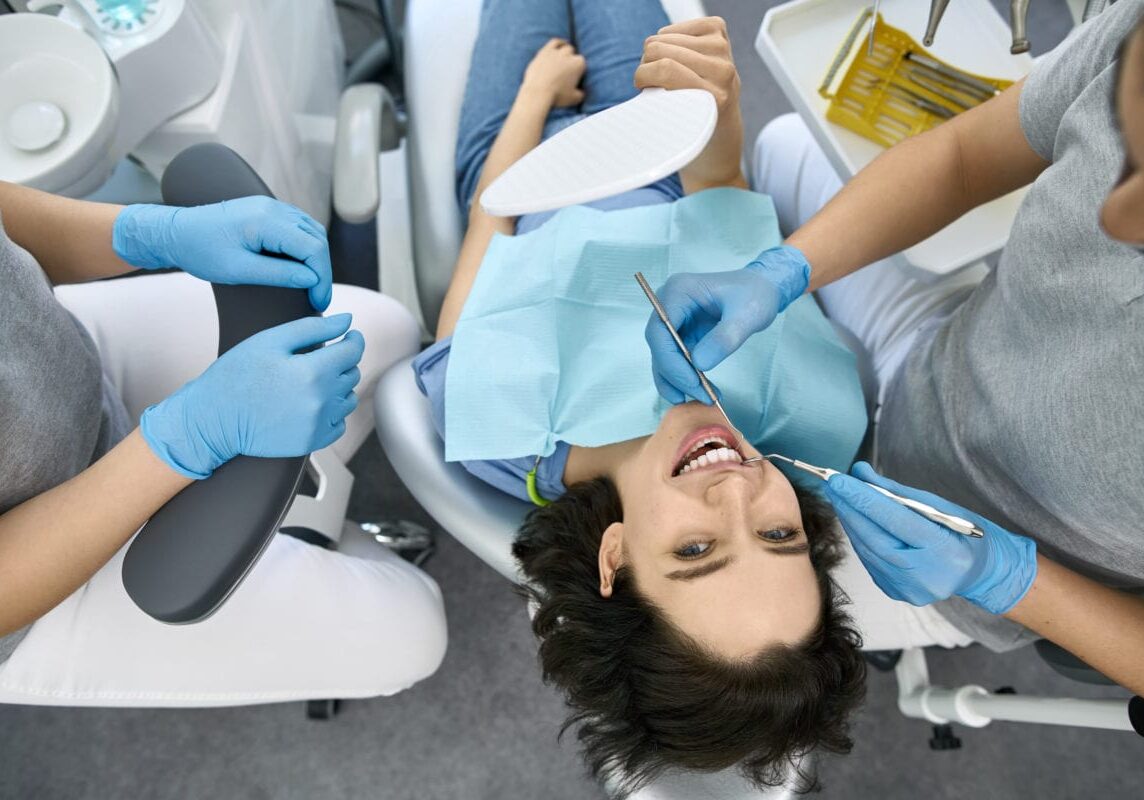 Glad girl in a patient's bib in a dental clinic. Dentist in latex blue gloves is examining her teeth with a help of a dental probe and a mirror. His assistant sits against him. Top view.